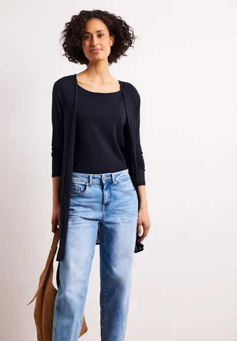 Een vrouw met krullend haar draagt een zwarte top, een vest, een lichtblauwe spijkerbroek en een bruine tas tegen een witte achtergrond.