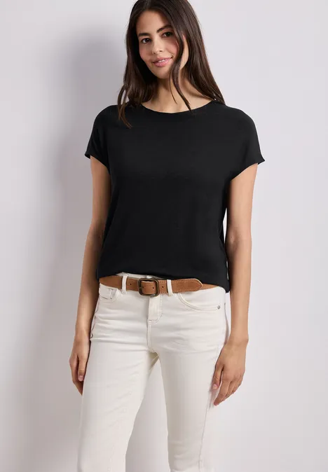 Frau in schwarzem T-Shirt, weißer Jeans und braunem Gürtel vor hellem Hintergrund.