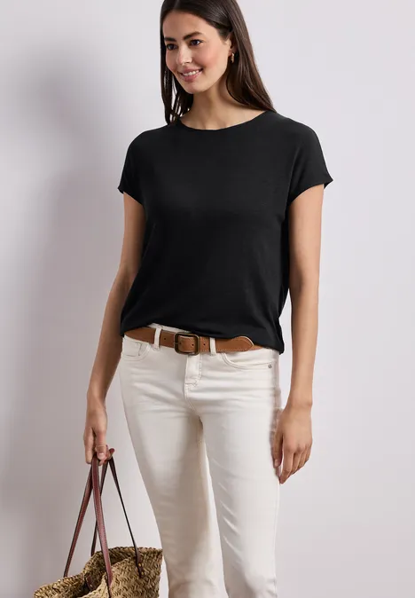 Eine Frau trägt ein schwarzes T-Shirt, weiße Hose, braunen Gürtel und hält eine Basttasche.