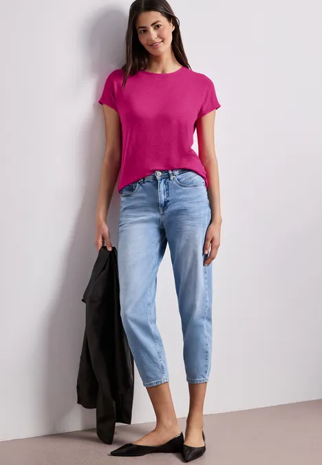 Frau in pinkem T-Shirt und hellblauen Jeans, hält schwarze Jacke, trägt schwarze Ballerinas vor hellem Hintergrund.