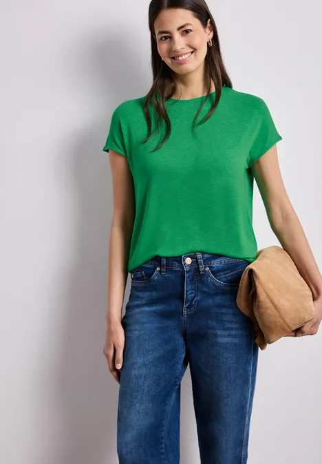 Eine Person trägt ein grünes T-Shirt, blaue Jeans und hält eine beige Tasche in der Hand.