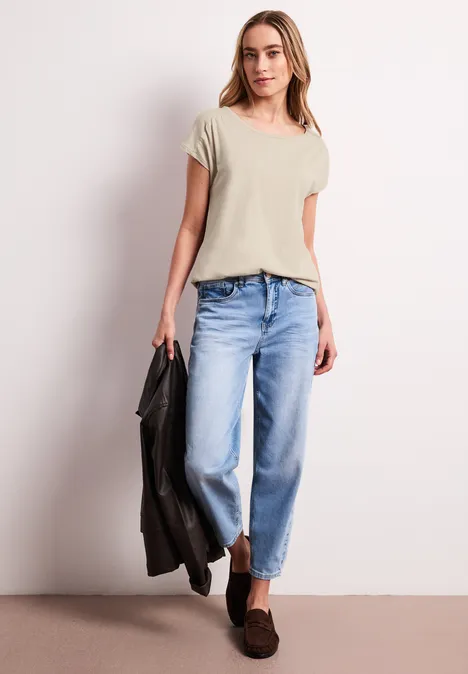 Vrouw met beige T-shirt, blauwe jeans, bruine loafers, houdt leren jack vast voor lichte achtergrond.