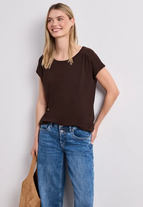 Een jonge vrouw draagt een eenvoudig bruin T-shirt, een blauwe spijkerbroek en houdt een tas vast.