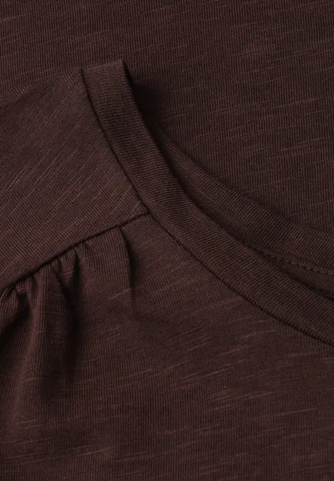 Close-up van een donkerbruin T-shirt met ronde hals en subtiele plooien bij de schouders.