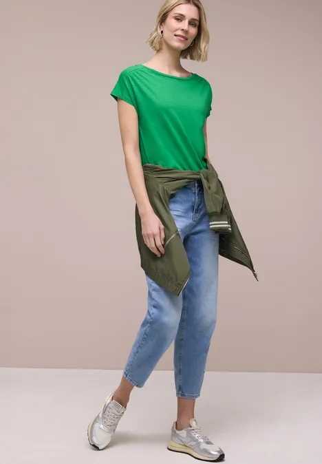 Vrouw in groen T-shirt, jeans en sneakers, met een jas om de taille gestrikt.