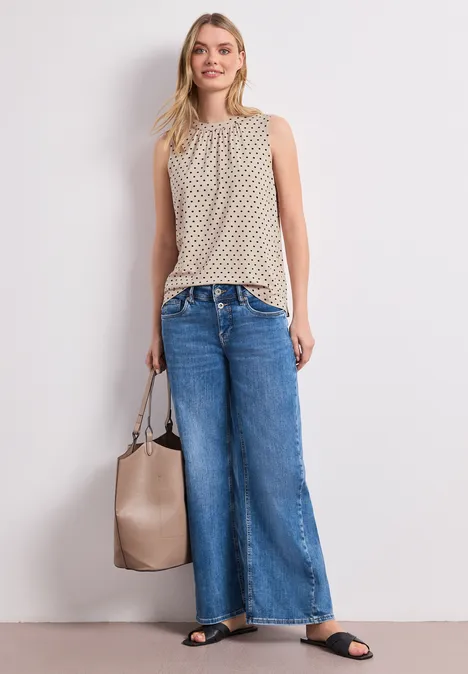 Frau in ärmellosem beigefarbenem Stricktop, weiten Jeans, schwarzen Sandalen und beiger Tasche.