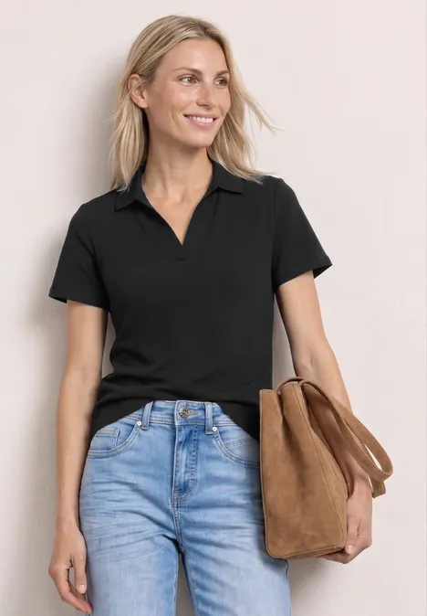 Frau mit schwarzem Poloshirt, blauen Jeans und brauner Tasche vor hellem Hintergrund.