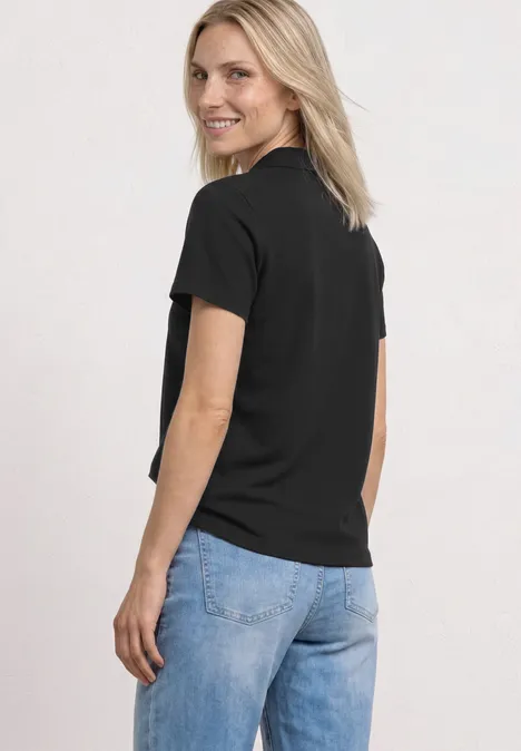 Eine Frau steht seitlich, trägt schwarzes T-Shirt und blaue Jeans vor weißem Hintergrund.