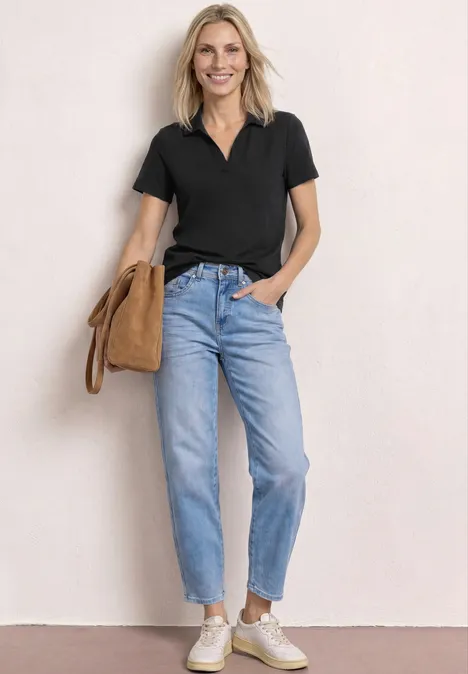 Frau in schwarzem Poloshirt, Jeans und weißen Sneakers, steht vor heller Wand mit beiger Schultertasche.