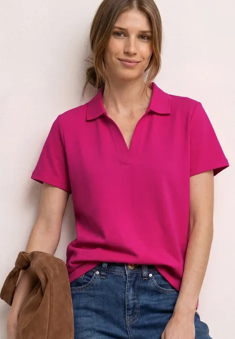 Vrouw in een roze poloshirt en blauwe jeans, met een bruine jas in de hand.
