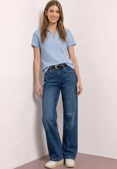 Een vrouw in lichtblauw poloshirt, blauwe jeans en witte sneakers staat nonchalant voor een neutrale muur.