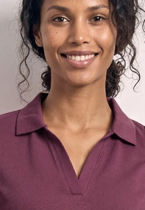 Een persoon met krullend haar draagt een paarse polo. Alleen het bovenlichaam en de hals zijn zichtbaar.