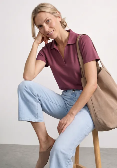 Iemand zit op een kruk, draagt een paars poloshirt, lichte jeans, beige schoenen en een tas.
