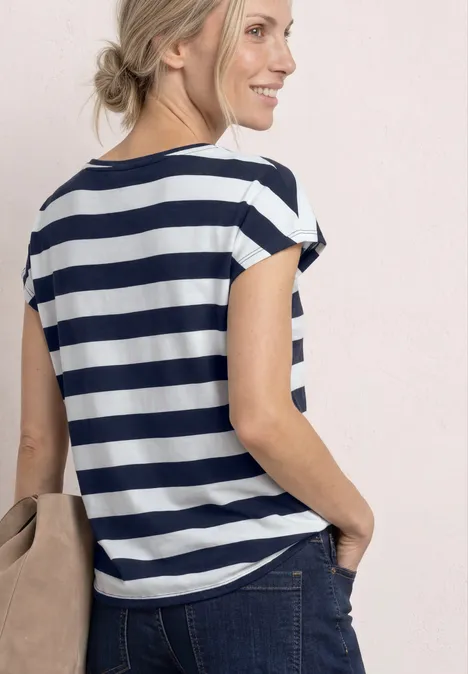 Vrouw van achteren met blauw-wit gestreept shirt, spijkerbroek en schoudertas voor lichte achtergrond.