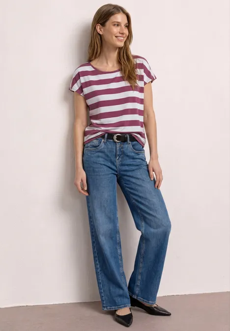 Shirt met korte mouwen, ronde hals en print dark clouded blush Vrouw met gestreept T-shirt, wijde spijkerbroek en platte zwarte schoenen, staand voor een lichte achtergrond.