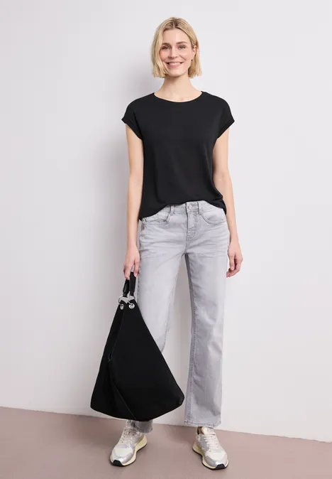 Person mit schwarzem T-Shirt, grauen Jeans, weißen Sneakern und einer großen schwarzen Tasche.