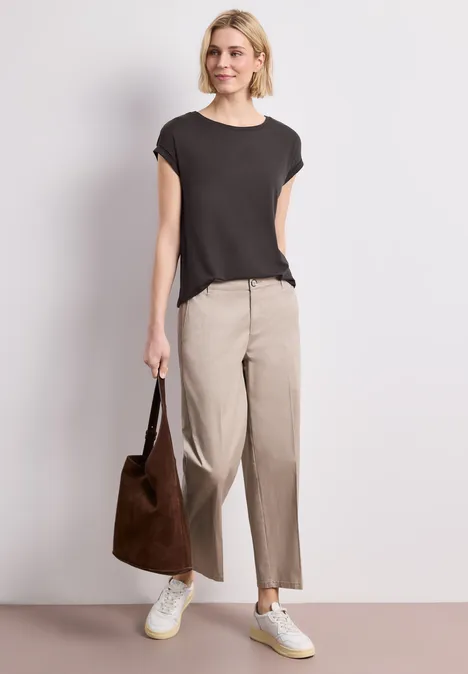 Eine Person trägt ein schwarzes T-Shirt, beige Hosen, weiße Sneaker und hält eine braune Tasche.