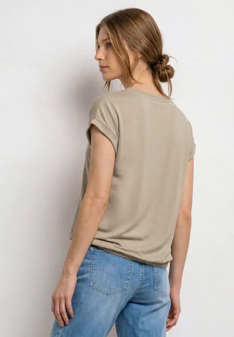 Frau in beigem T-Shirt und Jeans steht mit dem Rücken zur Kamera, die Haare zu einem niedrigen Dutt gebunden.