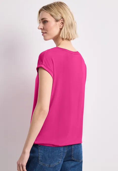 Frau von hinten in einem pinken Kurzarmshirt und blauen Jeans vor weißem Hintergrund.