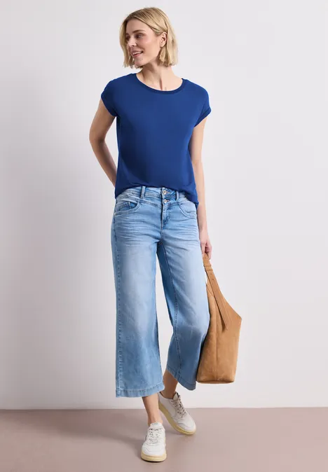 Frau in blauem T-Shirt und weiten Jeans mit weißen Sneakern und brauner Tasche.