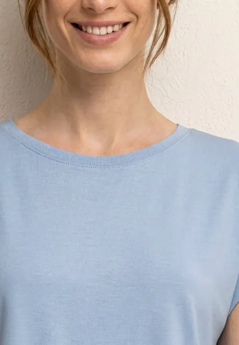 Eine lächelnde Frau in hellblauem T-Shirt vor neutraler Wand.