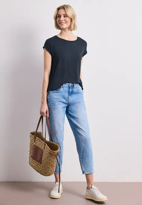 Frau in schwarzem T-Shirt, blauen Jeans und Sneakern, hält eine geflochtene Strohtasche.