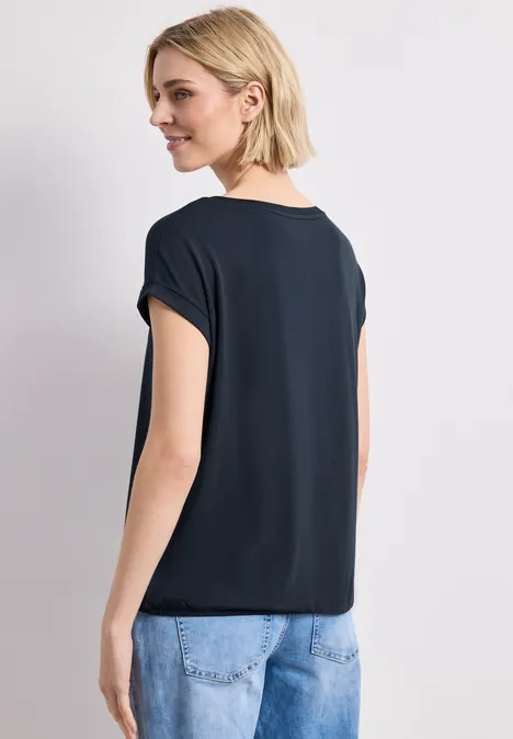 Eine Frau mit blondem Haar, von hinten gesehen, trägt ein schwarzes T-Shirt und blaue Jeans.