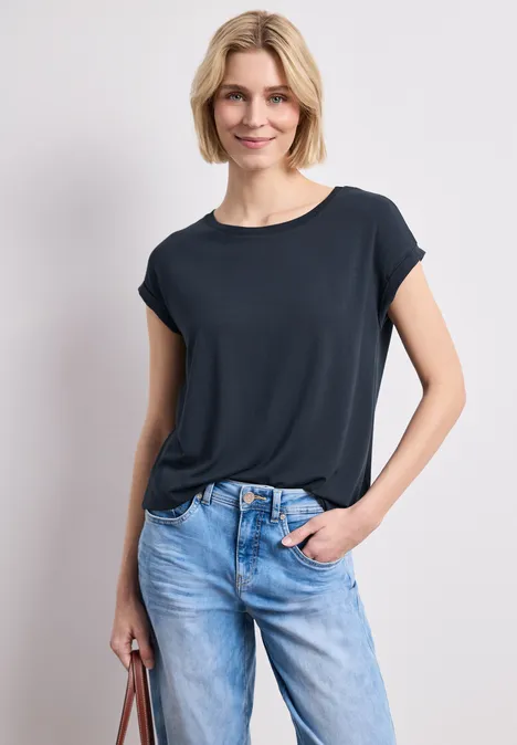Frau trägt dunkelblaues T-Shirt und helle Jeans, hält eine Handtasche.