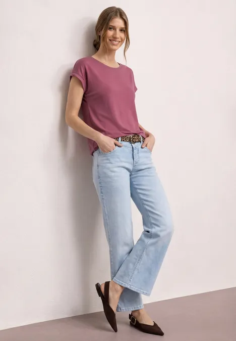 Eine Frau in pinkem T-Shirt, hellblauen Jeans und braunen Slippern lehnt lässig an einer hellen Wand.