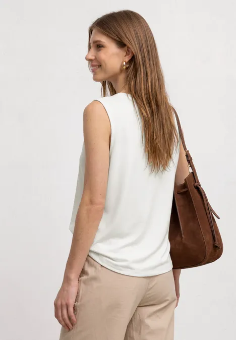 Frau mit weißem, ärmellosem Top und beigen Hosen, trägt eine braune Umhängetasche, blickt zur Seite.