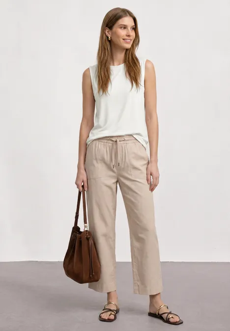 Eine Frau trägt ein weißes Top, beige Hosen und Sandalen und hält eine braune Handtasche.