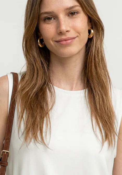 Frau mit langem braunem Haar in weißem ärmellosem Top und brauner Schultertasche.