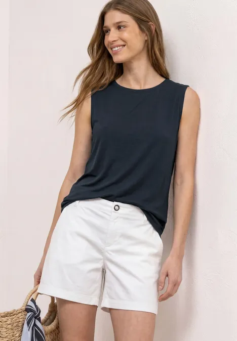 Frau trägt ärmelloses dunkelblaues Top, weiße Shorts und hält eine Strohtasche.
