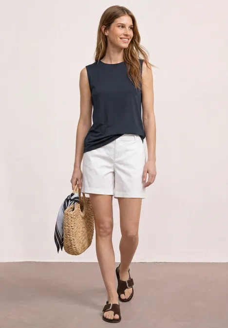 Frau in dunkelblauem Top, weißen Shorts, trägt Korbtasche und schwarze Sandalen vor neutralem Hintergrund.