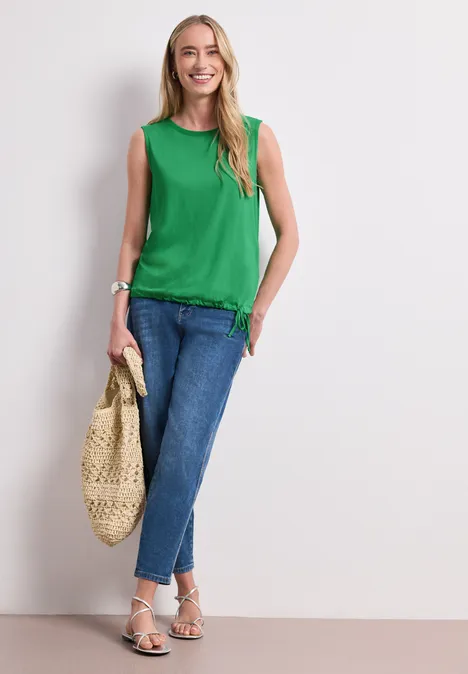 Top im U-Boot-Ausschnitt ever green Eine Frau trägt grüne Bluse, blaue Jeans, Sandalen und hält eine geflochtene Tasche.