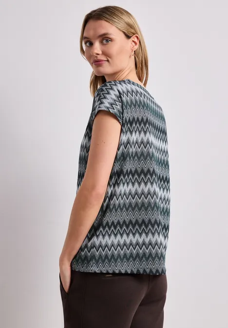 Femme vue de dos en haut à manches courtes zigzag gris-vert et noir, pantalon foncé.