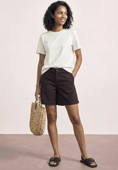 Vrouw in wit T-shirt, zwarte shorts en sandalen, met een rieten tas in haar hand.
