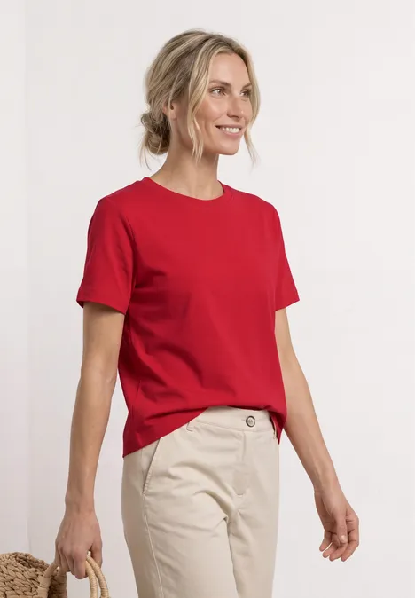 Persoon in rood T-shirt en beige broek voor een witte achtergrond, gezicht onscherp.
