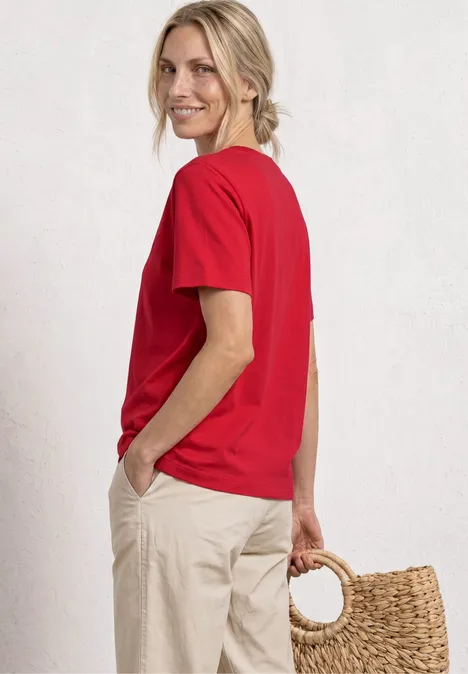 Vrouw op de foto van achter in rood T-shirt en beige broek, met gevlochten mand, lichte achtergrond