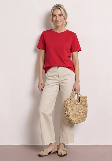 Persoon in rood T-shirt, beige broek en sandalen met een gevlochten mandtas voor een lichte achtergrond.