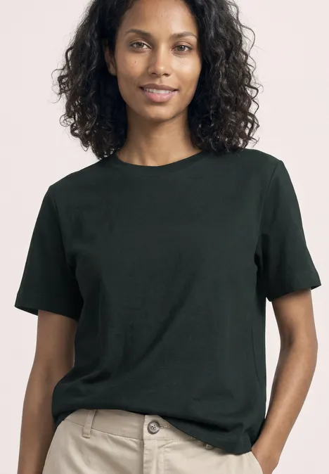 Een vrouw met krullend haar draagt een simpel zwart T-shirt en een lichte broek.