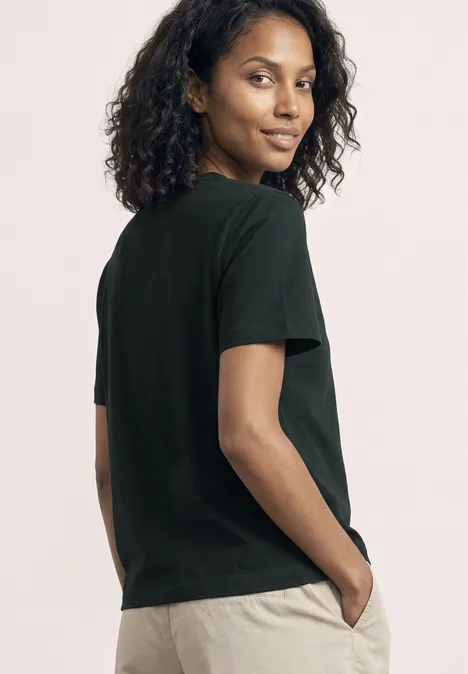 Van achteren gefotografeerde vrouw in zwart t-shirt en beige broek, handen in de zakken.