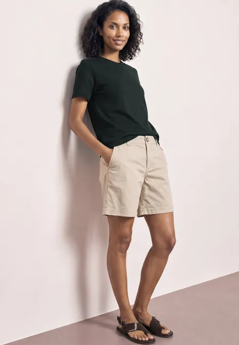 Iemand leunt tegen een muur, draagt een zwart t-shirt, beige short en zwarte sandalen.
