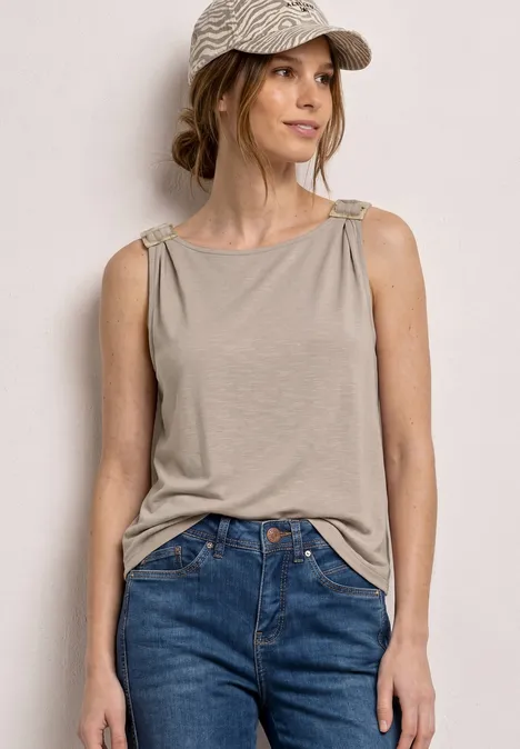 Vrouw in beige top met geknoopte bandjes, blauwe jeans en pet voor een lichte achtergrond.