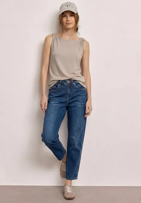 Een persoon staat voor een lichte muur met een beige tanktop, blauwe jeans en bijpassende loafers.