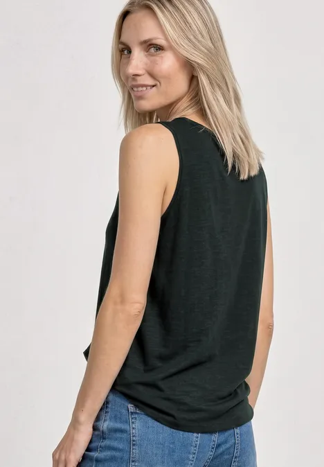 Een blonde vrouw in een zwarte mouwloze top en blauwe jeans, half zijwaarts gezien.