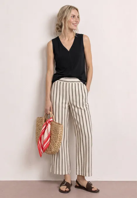 Frau in schwarzem Top, gestreifter Hose, braunen Sandalen und Korbtasche mit rotem Tuch vor hellem Hintergrund.