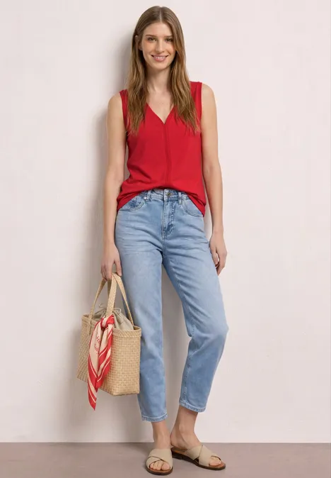 Vrouw in rode top, lichte spijkerbroek, strooien tas met sjaal, platte sandalen.