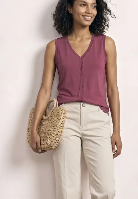 Eine Frau trägt ein ärmelloses, violettes Top mit V-Ausschnitt, helle Hose und hält eine runde Strohtasche.