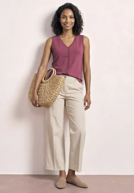 Frau mit lockigem Haar trägt burgunderfarbenes Top, beige Culottes, Basttasche und flache Schuhe.